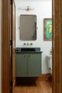 Small bathroom with floating green vanity, black stone countertop, wall-mounted mirror, brass fixtures, and hardwood flooring.