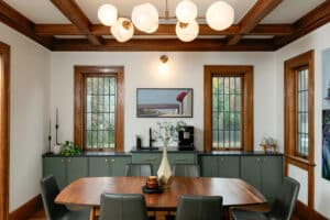 Renovated dining room featuring custom green built-in cabinetry, restored wood trim, coffered ceiling beams, modern brass lighting, and large windows bringing in natural light.