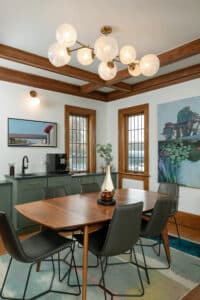 Finished dining room remodel with stained wood trim, sage green cabinets, brass hardware, wood table, and leather chairs with an abstract light fixture above
