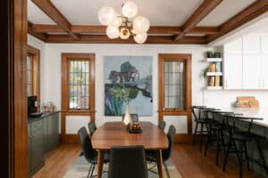 Finished dining room remodel with stained wood trim, sage green cabinets, brass hardware, wood table, and leather chairs with an abstract light fixture above. Kitchen peninsula on right side with blacks stools